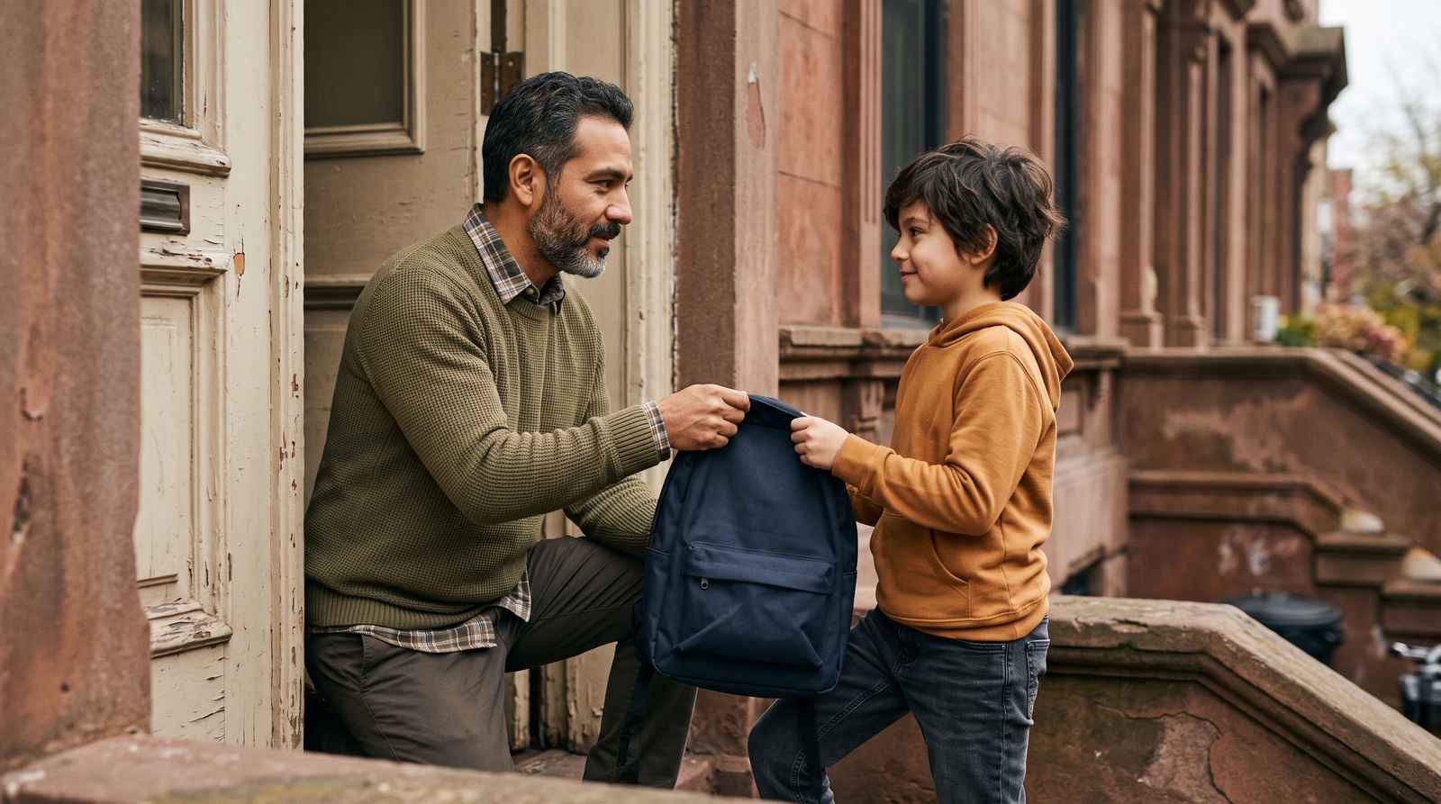 A father calmly hands his son a school backpack at the front door — everyday co-parenting.